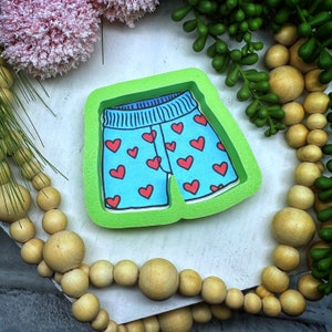 May include: Green silicone mold for baking or crafting. The mold is shaped like a pair of shorts with a blue and red heart pattern.