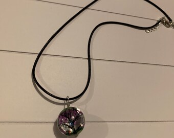 Puede incluir: Un collar de cordón negro con un colgante redondo y transparente. El colgante contiene un árbol en miniatura con detalles morados y verdes. El collar tiene un cierre plateado.