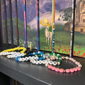 May include: Several beaded bracelets with white letter beads spelling out names and words. The bracelets are in various colors, including yellow, blue, green, and pink. The bracelets are displayed on a black shelf in front of a row of books.