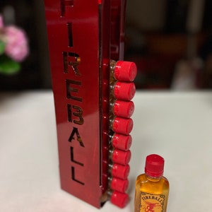 Pu&ograve; includere: Un espositore rosso per whisky alla cannella Fireball con la scritta "FIREBALL" ritagliata. L'espositore contiene diverse piccole bottiglie di whisky alla cannella Fireball, ciascuna con un tappo rosso. Una singola bottiglia di Fireball &egrave; accanto all'espositore.