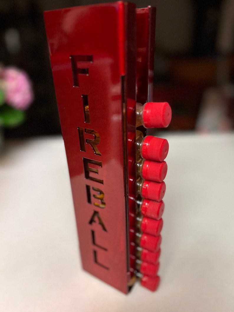 Pu&ograve; includere: Un dispenser per shot in metallo rosso con la scritta "FIREBALL" intagliata sul lato. Il dispenser contiene diverse bottigliette con tappi rossi. Lo sfondo &egrave; sfocato, con un accenno di composizione floreale.