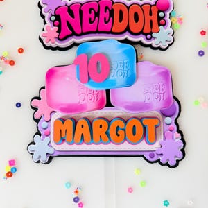 May include: A layered cake topper featuring the text NEEDOH in pink and orange, a blue square with the number 10 in pink, and the name MARGOT in orange. The design includes purple and blue splat shapes and a clear plastic stick at the base.