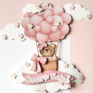 May include: A pink hot air balloon cake topper with a teddy bear in a basket. The balloon is made of pink spheres, with white clouds, stars, and a crescent moon. A pink banner reads "Amara Elaine".