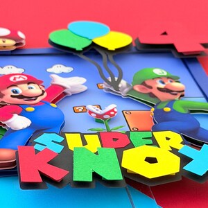 May include: A colorful pop-up card featuring Mario and Luigi characters. The card has a blue background with the words "SUPER KNOX" in large, multi-colored letters. Mario is wearing a red hat and blue overalls, while Luigi is in a green hat and overalls. Balloons and a mushroom are also visible.