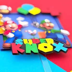 May include: A colorful 3D art piece featuring the text "SUPER KNOX" in bold, multi-colored letters. The artwork depicts cartoon characters Mario and Luigi, with a blue background and red, blue, and yellow accents. The overall theme is video game-related.