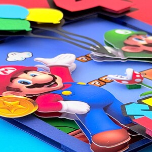 May include: A colorful, layered paper art piece featuring the character Mario from the Super Mario Bros. video game. Mario is in his signature red hat and blue overalls, with a gold coin and other game elements visible.