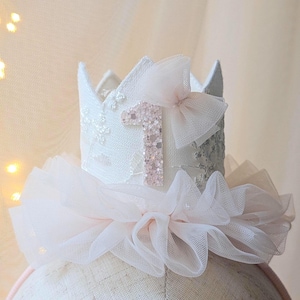 May include: A white fabric crown with a pink number 1 and a pink bow. The crown has a ruffled tulle collar and is decorated with lace. A festive accessory for a first birthday celebration.