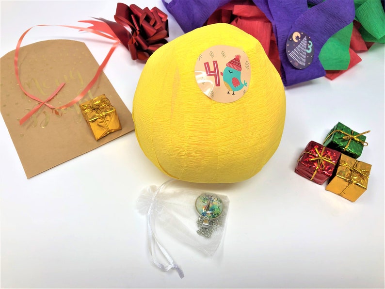 A large surprise ball rests on a white backdrop with Christmas decorations. The outer layer of the ball is yellow with a sticker that says #4. It marks the fourth day of an advent calendar.