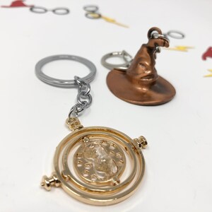 Close up of two keychains against a white backdrop. One is a golden time turner, while the other is a wizard or witch hat with a surly face.