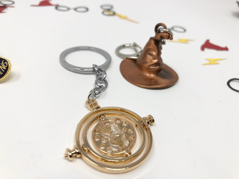 Close up of two keychains against a white backdrop. One is a golden time turner, while the other is a wizard or witch hat with a surly face.