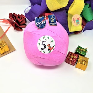 A large surprise ball rests on a white backdrop with Christmas decorations. The outer layer of the ball is pink with a sticker that says #5. It marks the fifth day of an advent calendar.