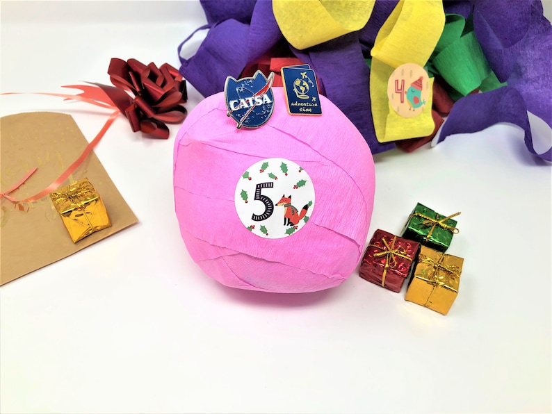 A large surprise ball rests on a white backdrop with Christmas decorations. The outer layer of the ball is pink with a sticker that says #5. It marks the fifth day of an advent calendar.