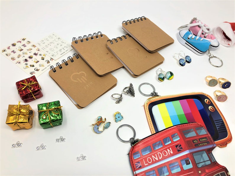 Various items are spread against a white backdrop. These items include pocket notebooks, keychains, rings, earrings, enamel pins, stickers, and more.