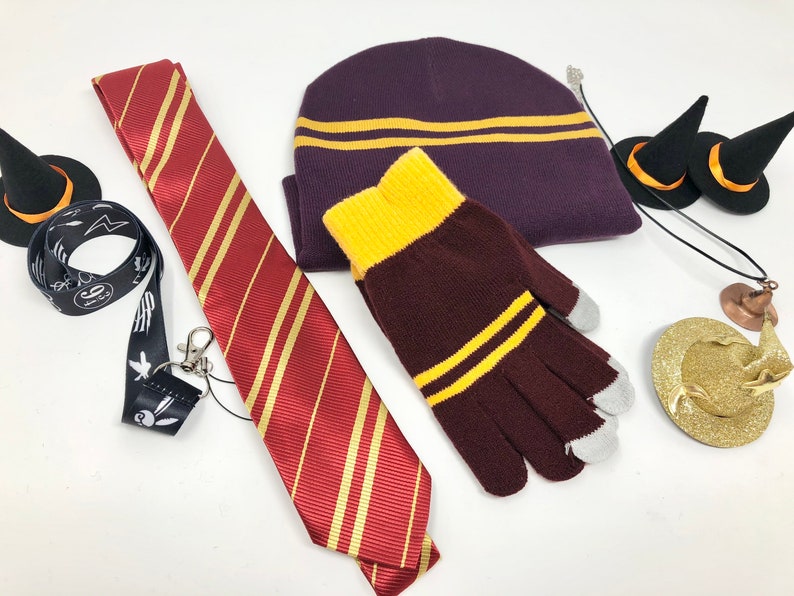 Various items are spread against a white backdrop. These include a lanyard, tie, beanie, gloves, necklace, and hair pin. The hair pin is topped with a small witch hat.