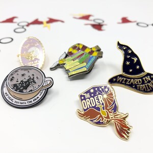 Close up of several enamel pins for book lovers. One is in the shape of a wizard hat and says Wizard in Training.