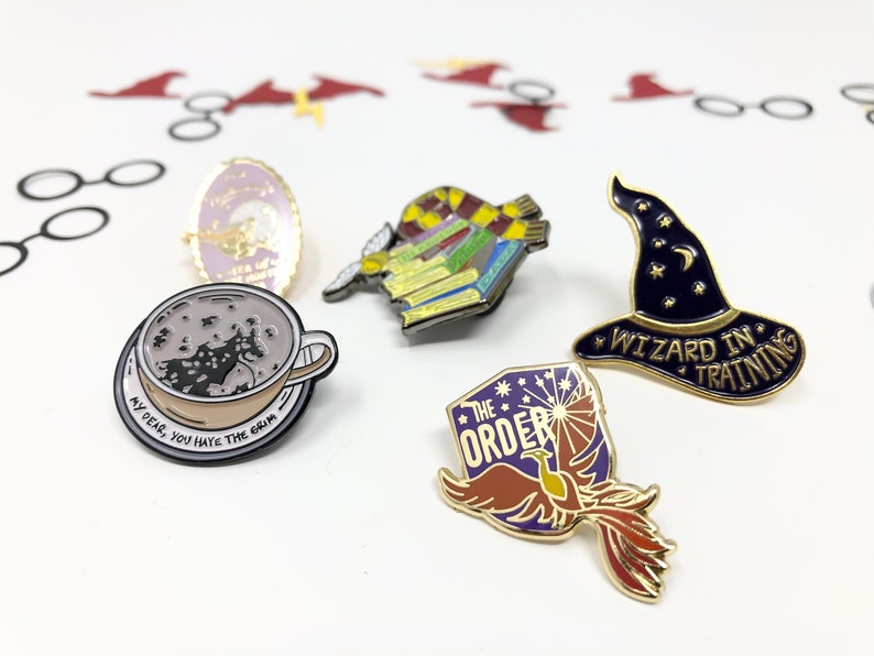 Close up of several enamel pins for book lovers. One is in the shape of a wizard hat and says Wizard in Training.