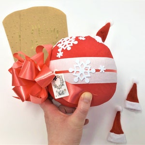 A large surprise ball rests on a white backdrop with Christmas decorations. The outer layer of the ball is red and white with a large bow. It is also decorated with a mini Santa hat.