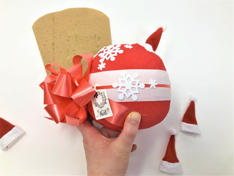 A large surprise ball rests on a white backdrop with Christmas decorations. The outer layer of the ball is red and white with a large bow. It is also decorated with a mini Santa hat.