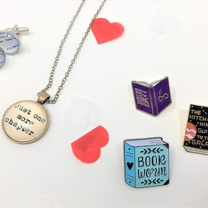 Close up of accessories against a white backdrop. These are themed pieces for book lovers. They include cuff links, a necklace, and enamel pins.