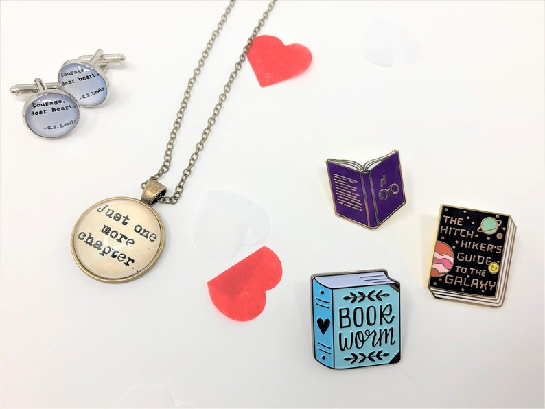 Close up of accessories against a white backdrop. These are themed pieces for book lovers. They include cuff links, a necklace, and enamel pins.