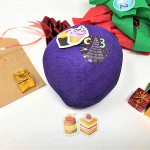 A large surprise ball rests on a white backdrop with Christmas decorations. The outer layer of the ball is purple with a sticker that says #3. It marks the third day of an advent calendar.