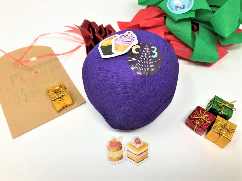 A large surprise ball rests on a white backdrop with Christmas decorations. The outer layer of the ball is purple with a sticker that says #3. It marks the third day of an advent calendar.