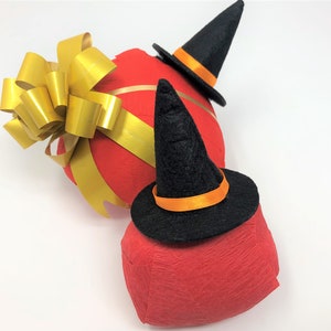 Red surprise balls against a white background. Each one has a red outer layer with a small witch hat on top. The larger one is also decorated with a golden bow.