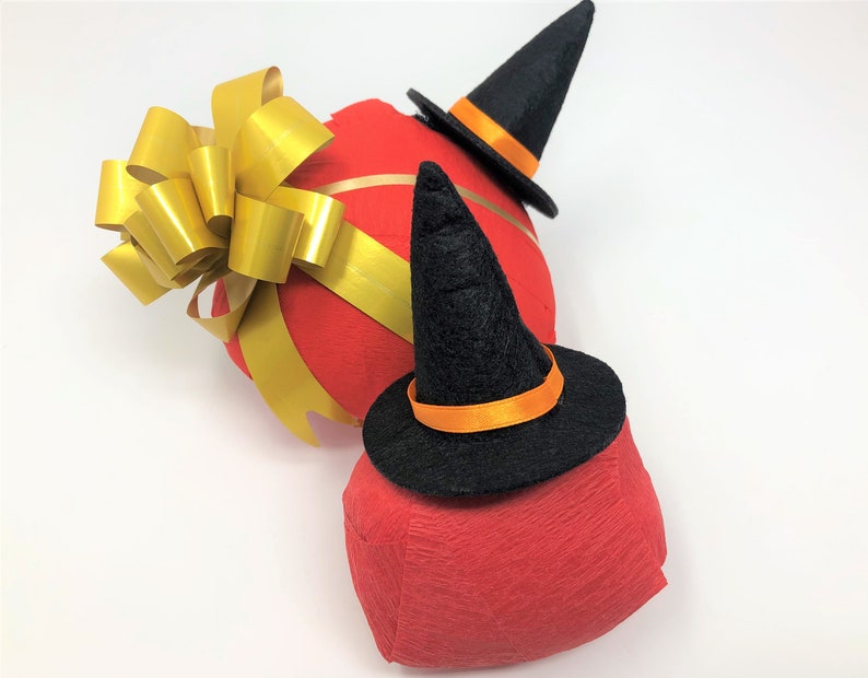 Red surprise balls against a white background. Each one has a red outer layer with a small witch hat on top. The larger one is also decorated with a golden bow.