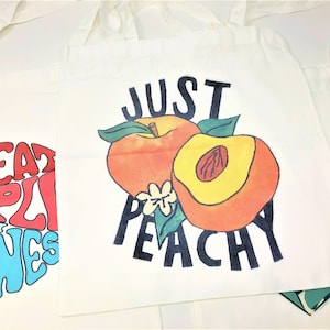 Several tote bags are spread across a white backdrop. One says stay peachy, while another says treat people with kindness. They are large and can be used for various purposes.