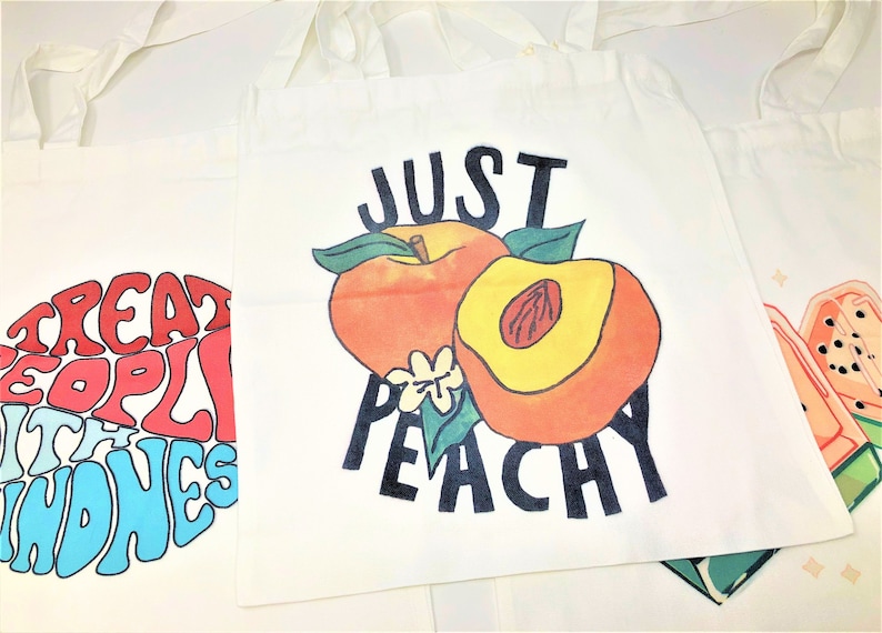 Several tote bags are spread across a white backdrop. One says stay peachy, while another says treat people with kindness. They are large and can be used for various purposes.