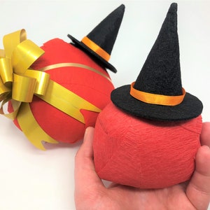 Hand holds a small surprise ball against a white background. Behind it lies a larger surprise ball. Each one has a red outer layer with a small witch hat on top. The larger size is also decorated with a golden bow.