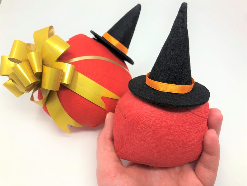 Hand holds a small surprise ball against a white background. Behind it lies a larger surprise ball. Each one has a red outer layer with a small witch hat on top. The larger size is also decorated with a golden bow.
