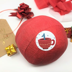 A large surprise ball rests on a white backdrop with Christmas decorations. The first layer of the ball is red with a sticker that says #1. It marks the first day of an advent calendar.
