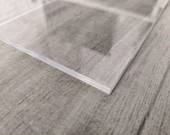 Puede incluir: Una lámina rectangular transparente de acrílico con una superficie lisa. Los bordes son limpios y bien definidos. La lámina está colocada sobre una superficie gris texturizada, creando un contraste entre el material transparente y el fondo.