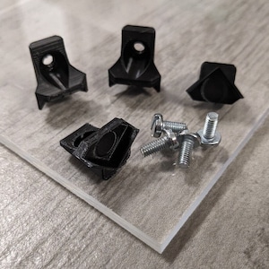Puede incluir: Cuatro soportes de esquina de plástico negro y seis tornillos plateados. Los soportes tienen forma triangular y están diseñados para conectar dos piezas de material en un ángulo de 90 grados.