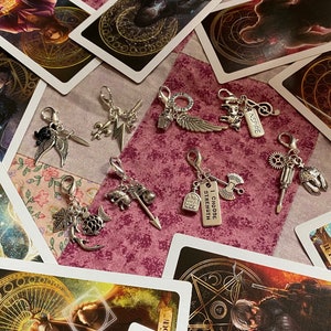 May include: A collection of silver charm pendants with various designs, including wings, a sword, a lightning bolt, a flower, a key, a bottle, an axe, a gear, a screwdriver, and a helmet. The charms are displayed on a pink and white patterned surface with tarot cards in the background.