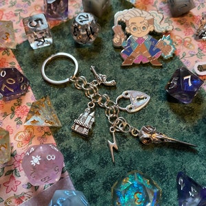 May include: A silver keychain with a variety of charms, including a castle, a lock and key, a lightning bolt, and a bird skull. The keychain is surrounded by iridescent dice with different numbers on them.