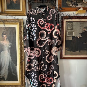 May include: Black long-sleeved dress featuring a snake print in red, white, and blue. The dress has a button-down front, a collared neckline, and a curved hemline. The dress is hanging on a black hanger.