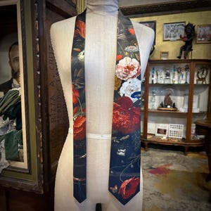 May include: A long necktie featuring a dark black background with a vibrant floral print. The pattern includes large red, white, and orange flowers, green foliage, and small butterflies. The tie is displayed on a neutral-coloured mannequin torso.