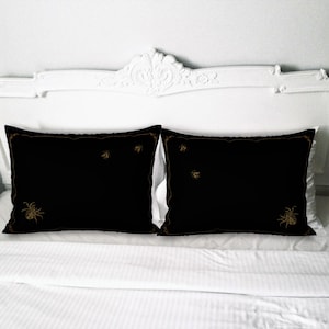 Könnte beinhalten: Zwei schwarze Kissenbezüge mit goldenen, gestickten Bienen und Spinnen. Die Kissenbezüge liegen auf einem weißen Bett mit einem weißen Kopfteil.