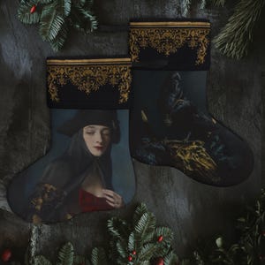 May include: Two Christmas stockings with a black background and gold trim. One stocking features a painting of a woman in a black dress and hat, the other features a painting of a raven on a skull.