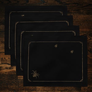 May include: Four black placemats with gold embroidered borders and insect and spider designs. The placemats are stacked on top of each other.