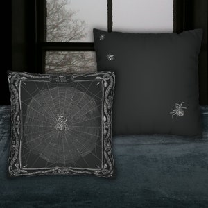May include: Two black throw pillows with white embroidered designs. One pillow features a spider web with a spider in the center and the text "Come into my parlor, said the spider to the fly". The other pillow has a spider and two flies.