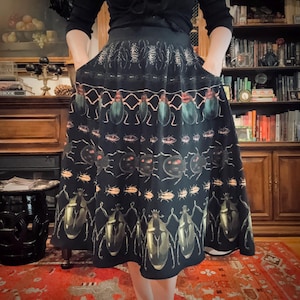 May include: Black skirt featuring a repeating pattern of beetles in green, gold, and brown. The skirt has pockets and a full shape. The background includes a wooden bookcase and a red patterned rug.