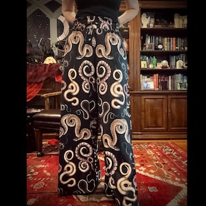 May include: Black palazzo pants with a repeating pattern of beige snakes. The high-waisted pants have a wide leg and a comfortable, flowing design. The snakes are stylized and create a unique, eye-catching print. The pants are made of a soft, lightweight fabric.