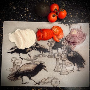 May include: A clear glass cutting board with a black crow design, topped with fresh ingredients. Includes a ripe avocado, tomatoes, sliced red onion, and a dollop of white cream. The board features crow illustrations and decorative chalices.