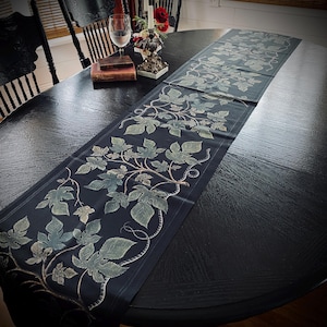 Op de afbeelding: Een zwarte tafelrunner met een herhalend patroon van groene bladeren en wijnstokken. De runner ligt op een zwarte houten tafel.
