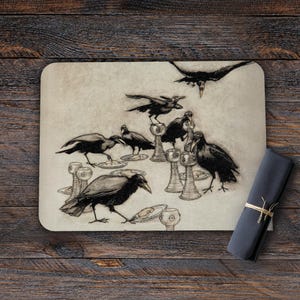 May include: A rectangular placemat with a detailed illustration of crows gathered around chalices and plates. The artwork is in black and white, with a vintage aesthetic. A rolled-up, dark gray napkin is tied with twine.