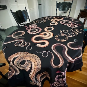 May include: Black oval tablecloth featuring a repeating pattern of various snake illustrations in shades of brown, tan, and white. The tablecloth is draped over a wooden table, with black chairs surrounding it.