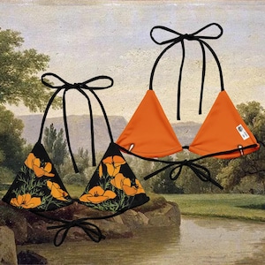May include: Two triangle bikini tops. One is solid orange, the other is black with a pattern of orange flowers. Both have black ties at the neck and back. The background is a landscape painting.